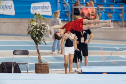 Young male gymnast in red leotard performs a graceful layout back flip mid-air at a Gymfed indoor competition.