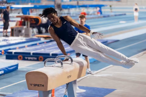 Young male gymnast performs a pommel horse routine, body fully extended in the air, intense focus on his face.