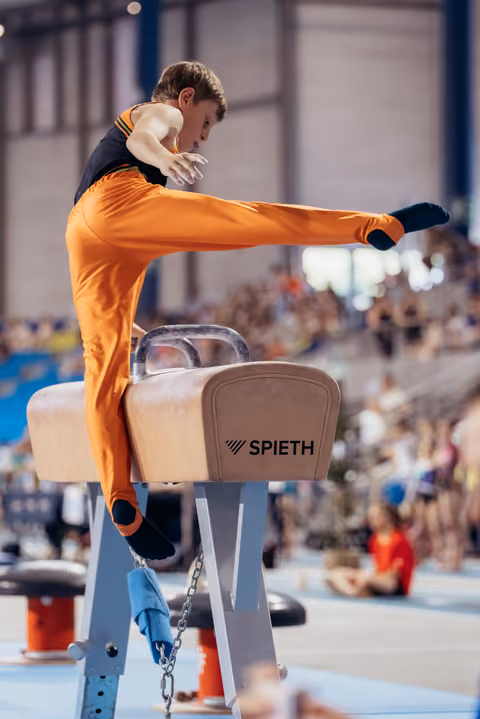 Young male gymnast in orange leotard performs a leg sweep on the pommel horse, focused and controlled.