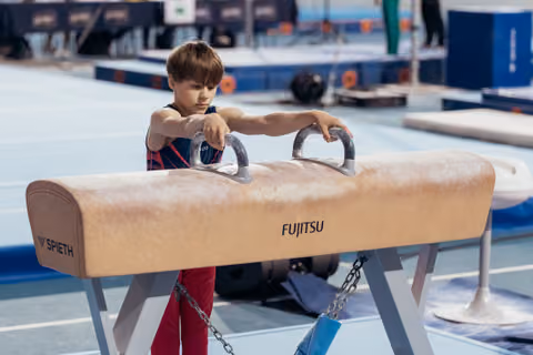 Young boy in gymnastics leotard grips pommel horse handles with intense focus in a blue competition gym.