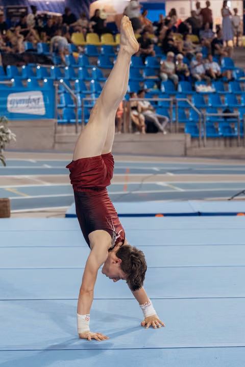 Young male gymnast performs a one-armed handstand on the floor, showing intense focus and strength during a competition.