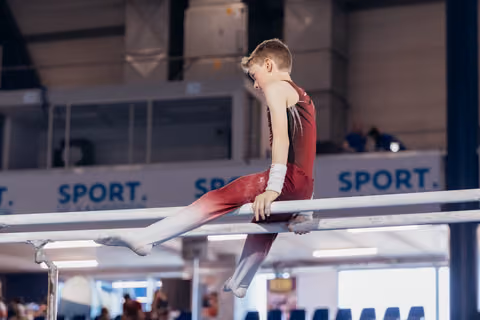 Young male gymnast in red leotard sits on parallel bars, looking down with focused concentration during a gymnastics meet.