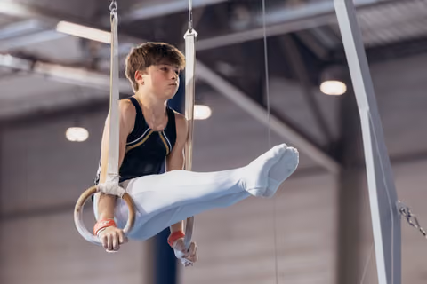Young male gymnast holds an L-sit on still rings, expression focused and determined, in an indoor gymnastics hall.