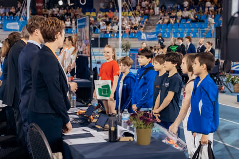 Young gymnasts in blue jackets line up attentively before officials at a registration table inside a packed sports arena.