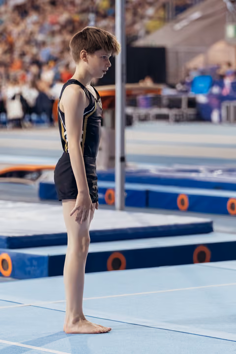 Young male gymnast stands barefoot on the floor mat, expression serious and concentrated, preparing for his routine.