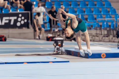 Young female gymnast mid-backflip, body fully inverted, arms extended, performing on floor exercise in competition arena.