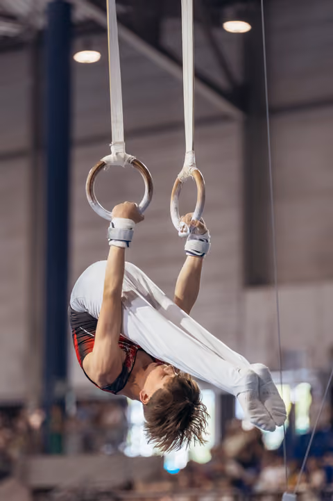 Young male gymnast performs an inverted hang on still rings, body arched, face tense with effort, in a packed arena.