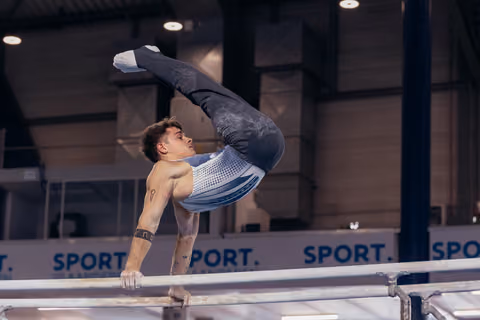 Male gymnast performs a dramatic pike hold on parallel bars, body arched with legs extended upward in a competition hall.