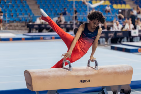 Young male gymnast performs a precise pommel horse routine, legs split wide, focused expression in a competition arena.