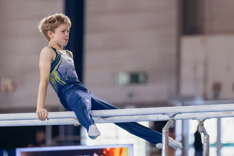 Focused young male gymnast seated on parallel bars, legs extended, wearing blue and green leotard in indoor gymnastics hall.