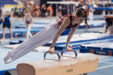 Focused young male gymnast performs a planche on a Fujitsu pommel horse during a gymnastics competition.