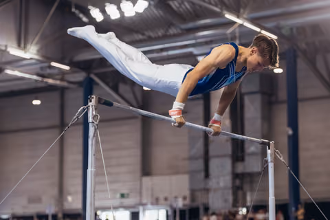Young male gymnast performs a tuck hold on the horizontal bar, body parallel, focused expression in a large sports hall.