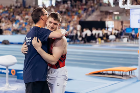 Young male gymnast hugs his coach on the competition floor, both sharing a warm, emotional moment of support.
