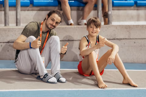 Young male gymnast and coach sit together on the mat, both smiling and giving thumbs up in a cheerful moment.