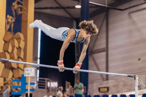 Young male gymnast performs a straddle hold on the horizontal bar, face intense with concentration, in a competition hall.