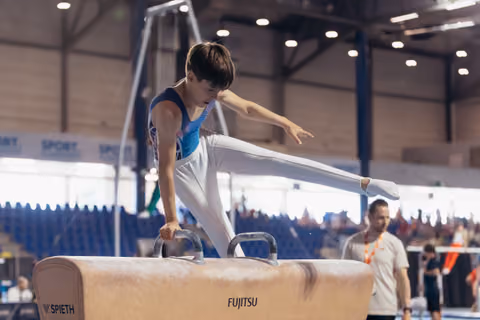Young male gymnast concentrates intensely on pommel horse, leg raised mid-routine, coach watching in background.