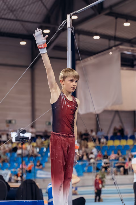 Young male gymnast raises hand confidently before high bar routine in a packed indoor gymnastics arena.