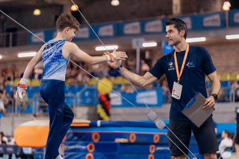 Young male gymnast shakes hands with smiling coach holding clipboard, sharing a proud moment on the competition floor.