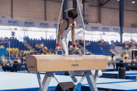 Young male gymnast concentrating intensely on pommel horse, leaning forward with red wristbands, in a packed arena.