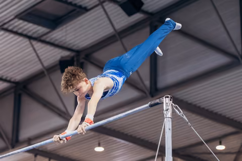 Young male gymnast performs a dynamic horizontal bar routine, body fully extended, expression intense with concentration.