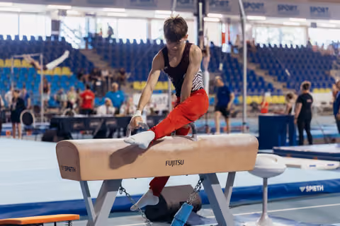 Young male gymnast leans intently over a Fujitsu-branded pommel horse, gripping pommels with focused concentration in a large arena.