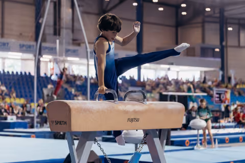 Young male gymnast performs on pommel horse, leg raised high, intense focus on face, in a packed competition arena.