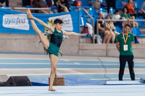 Young gymnast performs a standing split on floor exercise, arms extended, concentrated expression, with coach watching nearby.