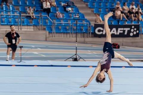 Young gymnast performs a one-handed cartwheel on the floor exercise mat during a competition, observed by a coach.