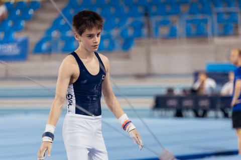 Young male gymnast stands focused and alert on the competition floor, wearing navy leotard and white trousers with wrist guards.