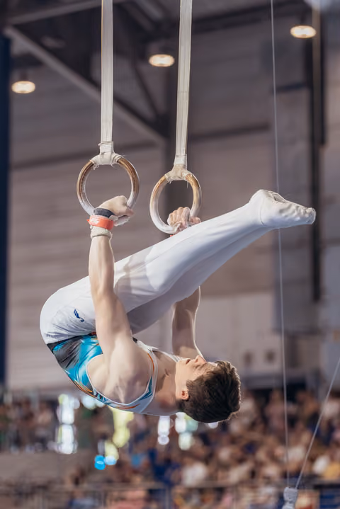 Young male gymnast performs an inverted tuck hold on still rings, showing intense concentration during a competition.