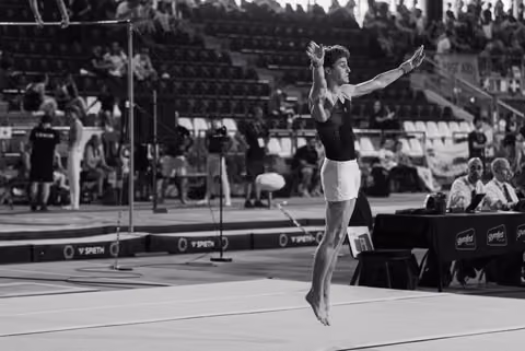 Male gymnast mid-air during floor exercise, arms gracefully extended, focused expression, judges seated nearby.
