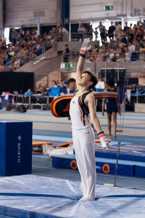 Young male gymnast stands tall on mat, one arm raised confidently toward the rings, wearing Belgian colors in a packed arena.