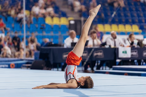 Young male gymnast performs a leg raise on floor, body arched gracefully, during a competition in a packed arena.