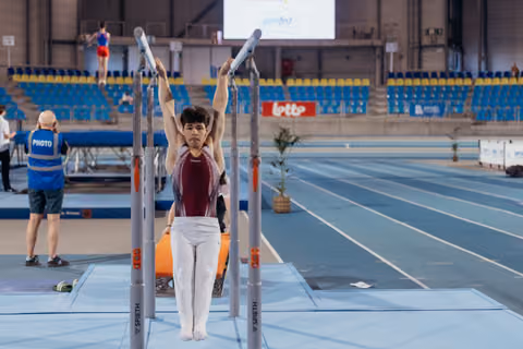 Young male gymnast hangs from parallel bars with focused expression, white pants and maroon leotard, indoor arena.