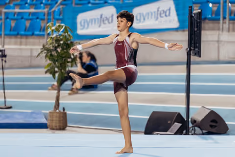 Young gymnast balances on one leg with arms extended, performing a floor routine with focused expression at Gymfed event.