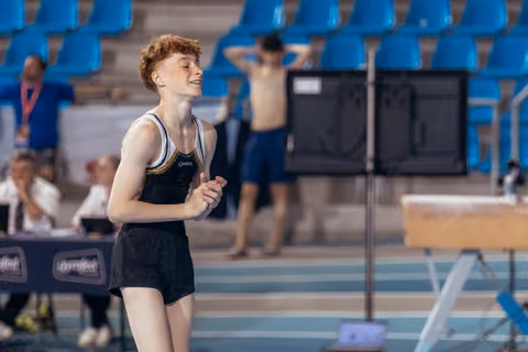 Young redheaded gymnast smiles with clasped hands after a routine, eyes closed, showing relief and joy at an indoor gymnastics meet.