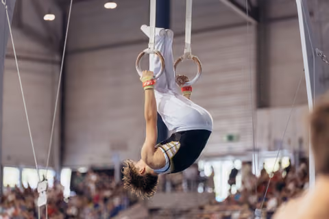 Young male gymnast performs an inverted hold on still rings, showing intense focus and strength in a packed arena.