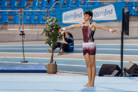 Young male gymnast stands barefoot on floor mat, arms wide open, composed expression, during a Gymfed indoor event.