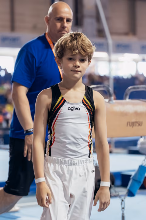 Young male gymnast stands composed and focused, wearing Agiva leotard, with coach standing behind him at gymnastics event.