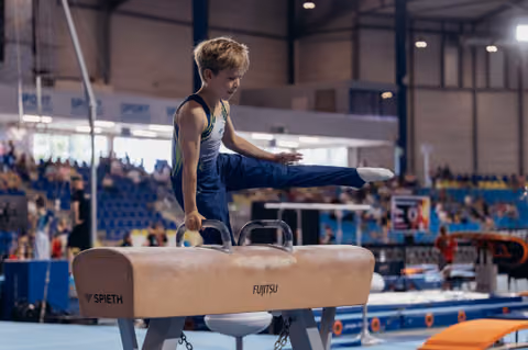 Focused young male gymnast performs on pommel horse, leg extended, in a busy competition arena.