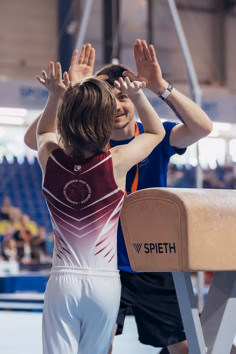 Young gymnast and smiling coach share an enthusiastic high-five beside a Spieth pommel horse in a gymnastics hall.