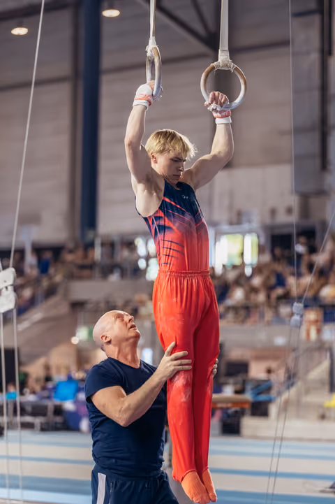 Young blond gymnast grips still rings with intense focus while coach spots his hips in a gymnasium.