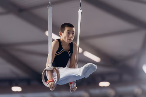 Young male gymnast holds a tucked position on still rings, expression focused and determined, inside a gymnasium.
