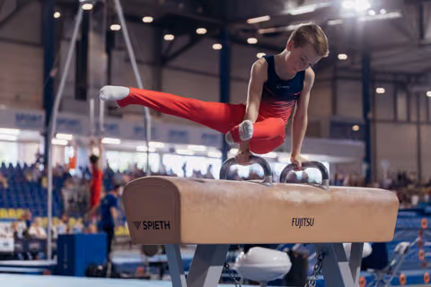 Young male gymnast performs focused pommel horse routine, legs extended, at an indoor gymnastics competition.