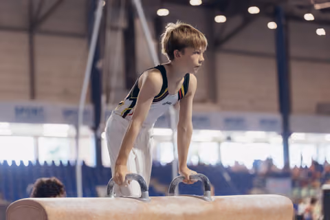 Young gymnast stands focused on pommel horse, gripping handles with determined expression in a sports hall.
