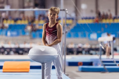 Young male gymnast in red Quatro leotard stands pensively at pommel horse, calm expression, in a large gymnastics arena.