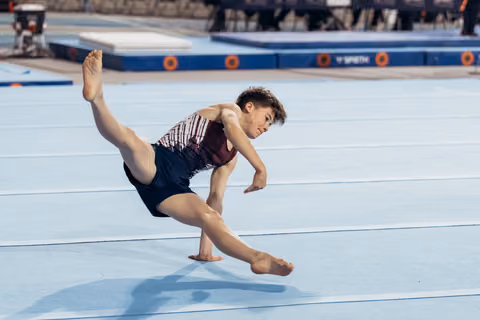 Young male gymnast mid-leap on floor exercise, body stretched low, intense focus on face during competition.