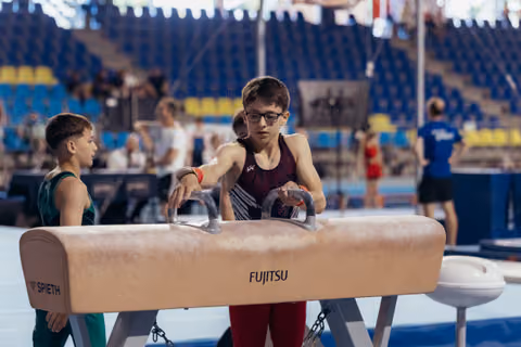 Young male gymnast with glasses grips pommel horse handles with focused concentration, teammate watches in background at indoor arena.