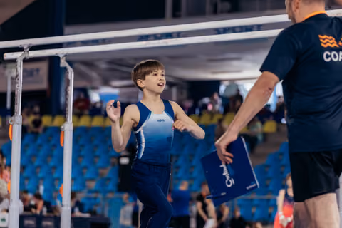 Young male gymnast walks toward coach with an eager expression, parallel bars behind him in a competition arena.