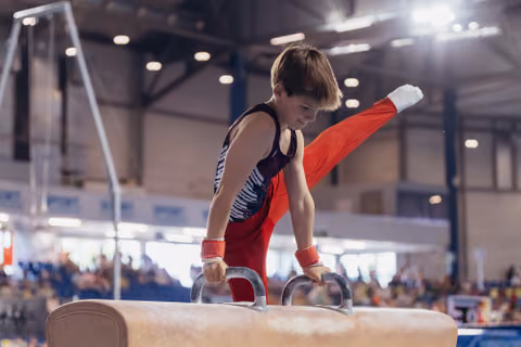 Young male gymnast performs on pommel horse, leg raised high, expression focused and determined, indoor arena setting.